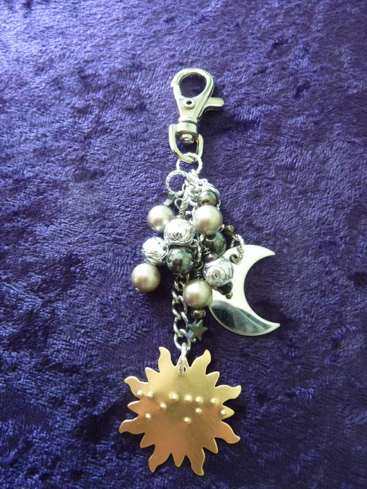 A purse charm with a sun, moon, and stars motif. The word "Shine" is written in braille